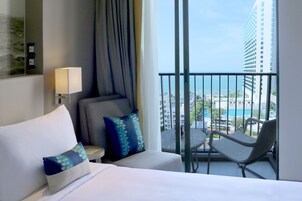 Habitación de hotel con cama, una silla, una mesa y balcón con vista a un edificio y una piscina.