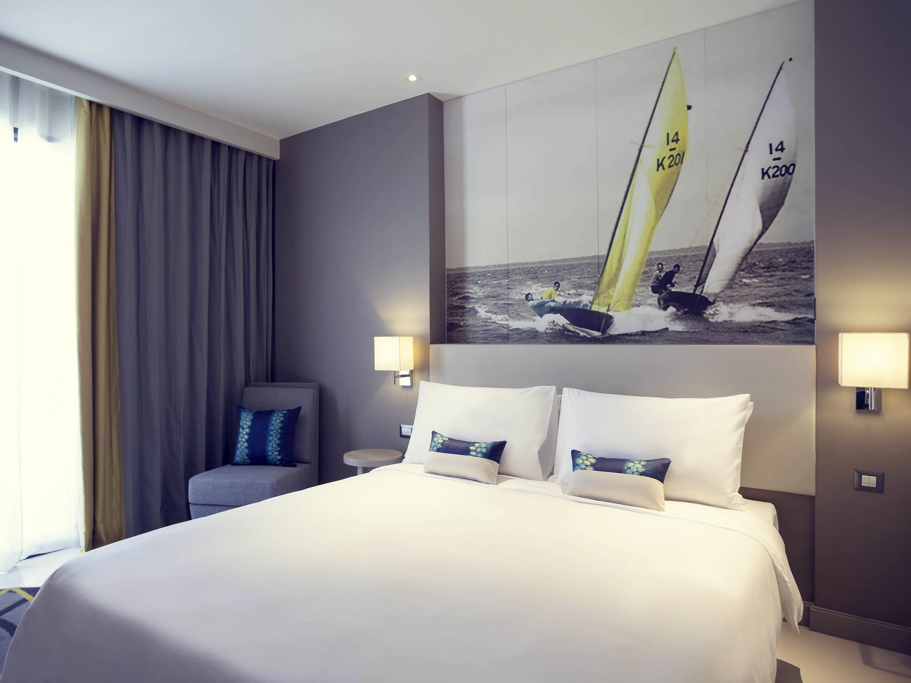 A hotel room with a large bed, two bedside lamps, a chair, a painting of sailboats, and a window with curtains.