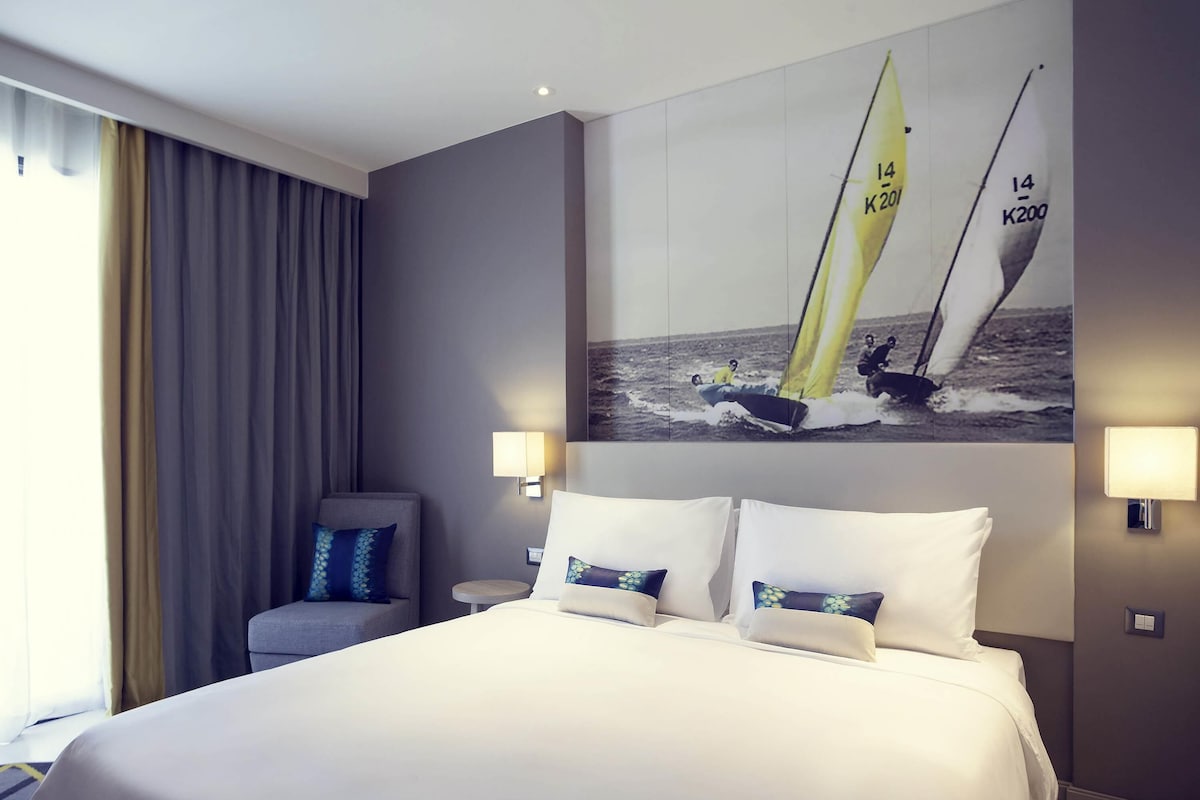 A hotel room with a large bed, two bedside lamps, a chair, a painting of sailboats, and a window with curtains.