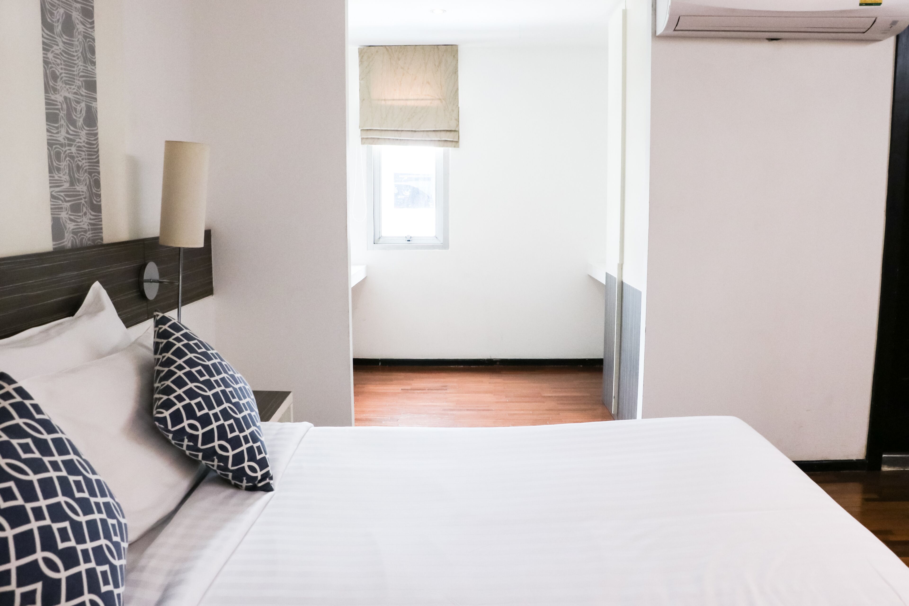 A hotel room with a bed, a nightstand, a lamp, a window, and a wall-mounted air conditioner.