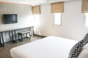 A modern hotel room with a large bed, a desk with two chairs, a flat-screen TV mounted on the wall, and two windows with beige curtains.