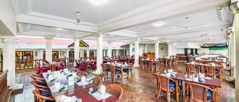 A spacious restaurant with multiple dining tables and chairs, white ceiling fans, and a view of a colorful building in the background.