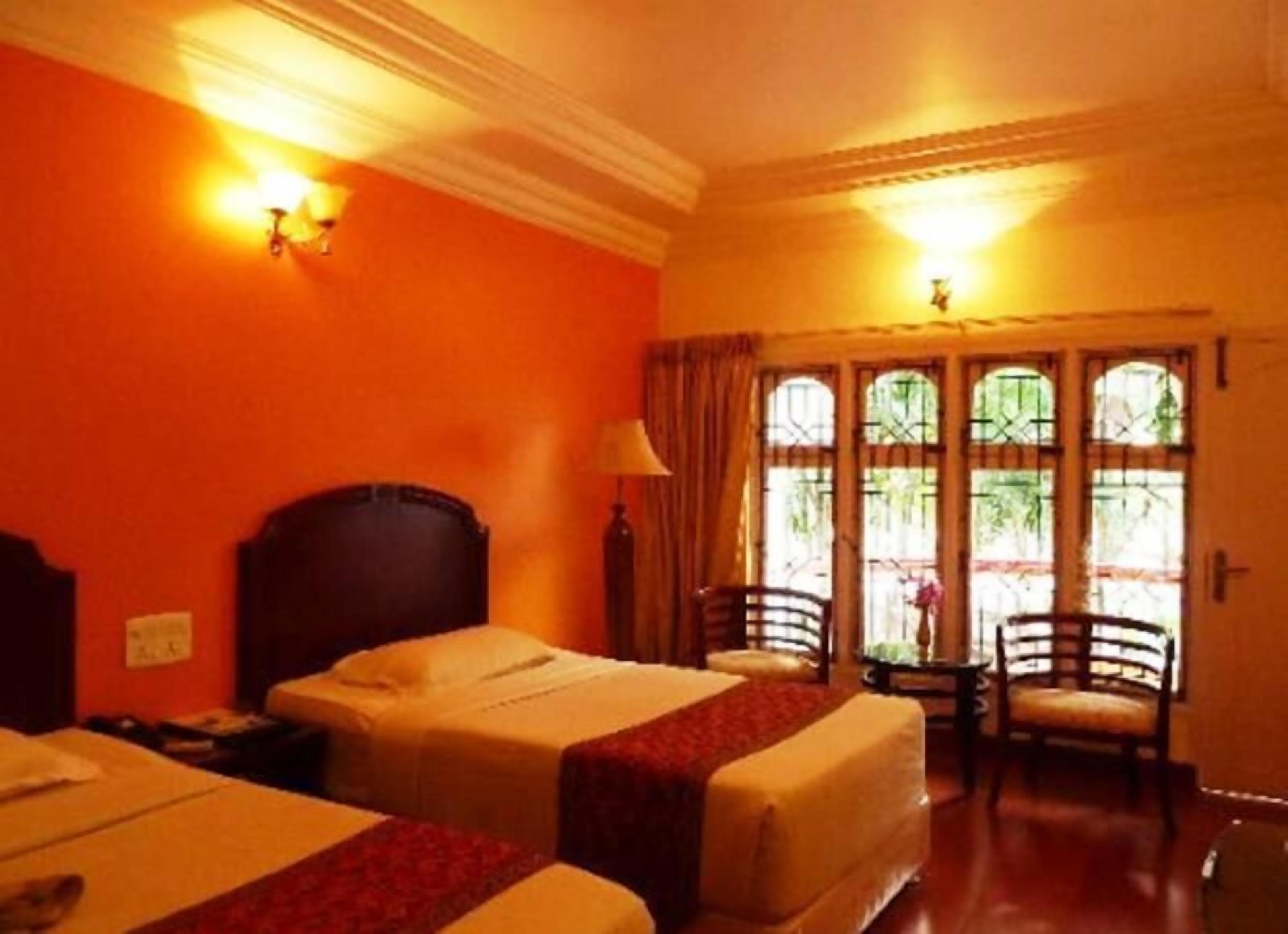 A hotel room with two beds, a wooden headboard, a nightstand, a lamp, and a view of the outdoors through a window with bars.