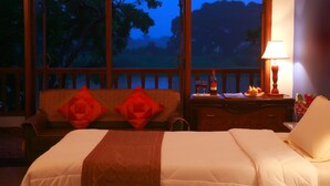 Habitación de hotel con cama, mesita de noche con lámpara y vista a árboles a través de amplios ventanales.