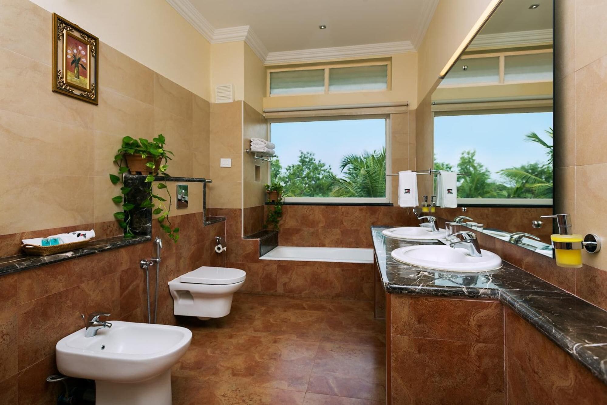 A modern bathroom with two sinks, a bidet, and a toilet. There are plants, a large mirror, and a window providing a view of greenery.