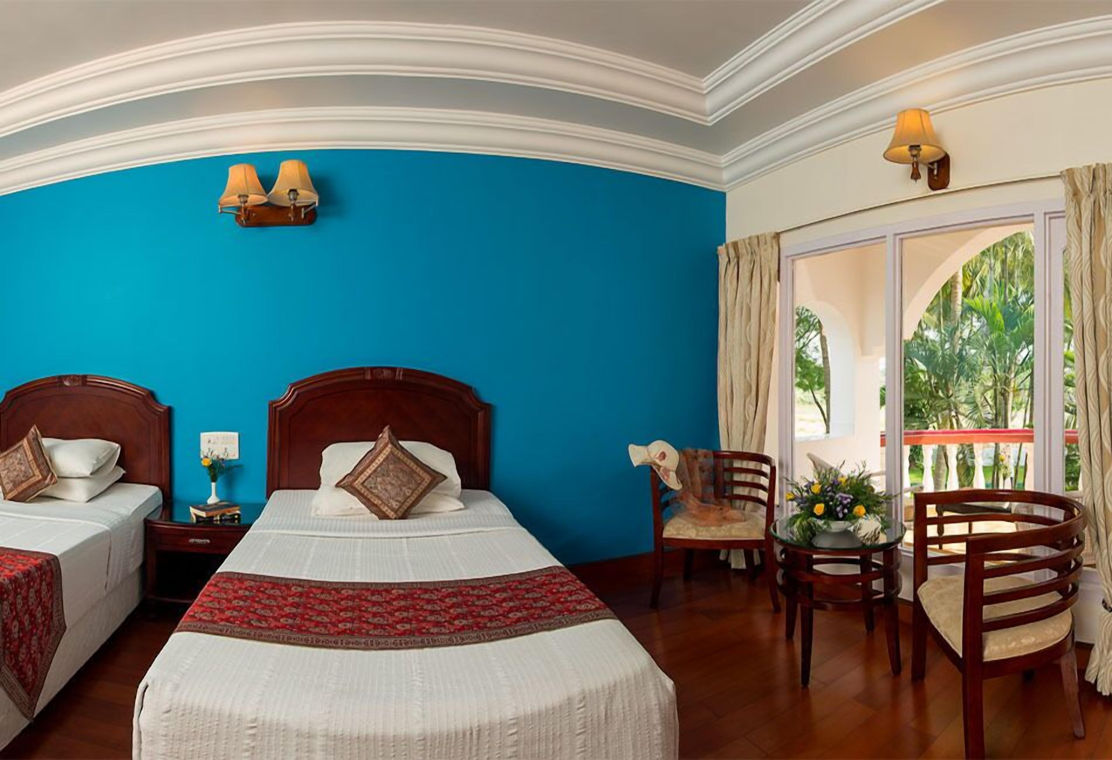 Une chambre d’hôtel avec deux lits, une table à manger avec des chaises, une fenêtre avec des rideaux et une vue sur la verdure environnante.