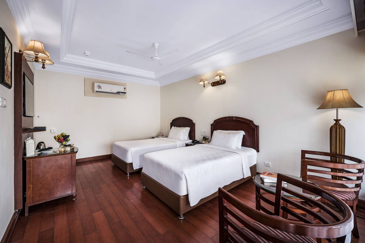 Ein Hotelzimmer mit zwei Einzelbetten, einem Holztisch, einem Stuhl, einer Lampe und einem Deckenventilator.