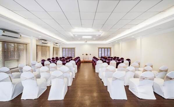 A conference room with rows of white chairs and tables, air conditioning units, and windows with blinds. - Great Trails River View Resort Thanjavur by GRT Hotels (Thanjavur)