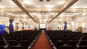 Un salón de conferencias con filas de sillas, un escenario al frente y banderas en las paredes.
