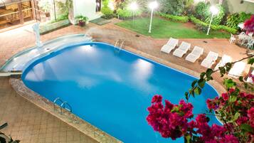 Une piscine intérieure à l’eau d’un bleu limpide, entourée d’une terrasse carrelée et d’un petit jardin agrémenté de plantes en pot et de quelques chaises.