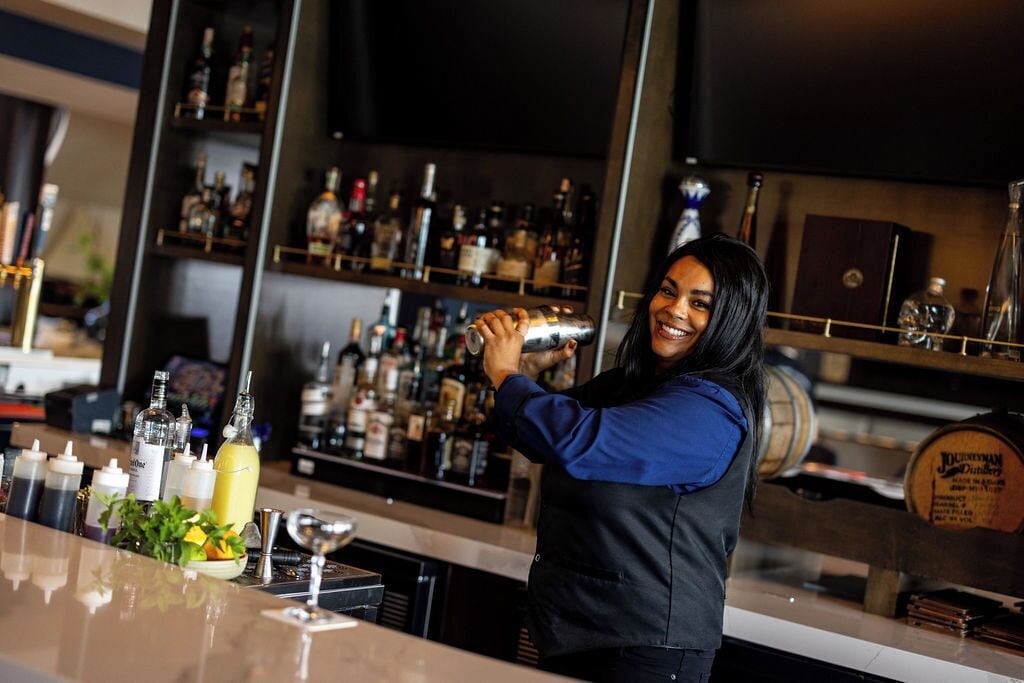 Un barman prépare un cocktail dans un bar où l’on aperçoit, derrière lui, des étagères remplies de bouteilles et un tonneau en bois.