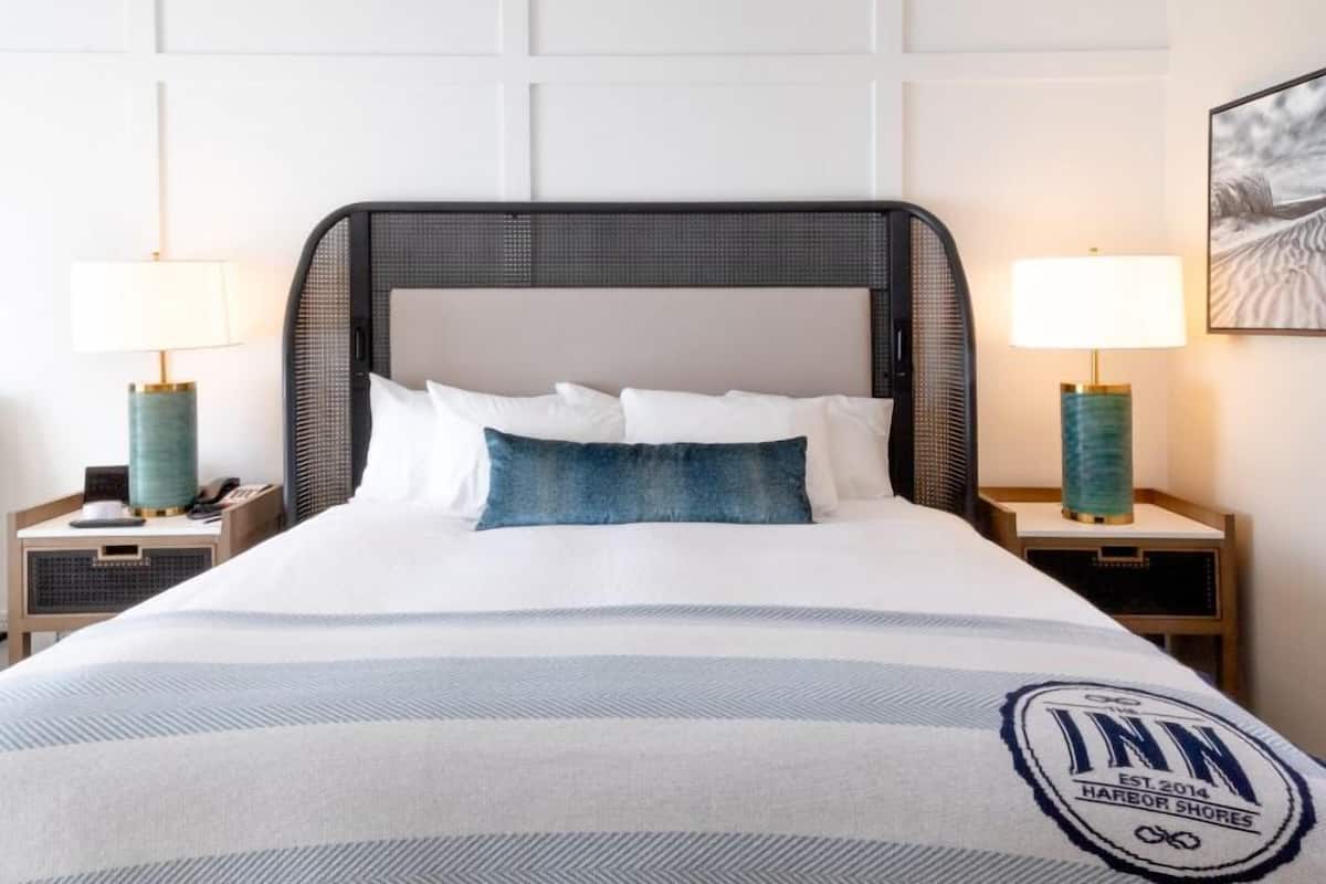 Ein Hotelzimmer mit einem großen Bett, zwei Nachttischen mit Lampen, einem gerahmten Bild an der Wand und einem Logo auf der Bettdecke.