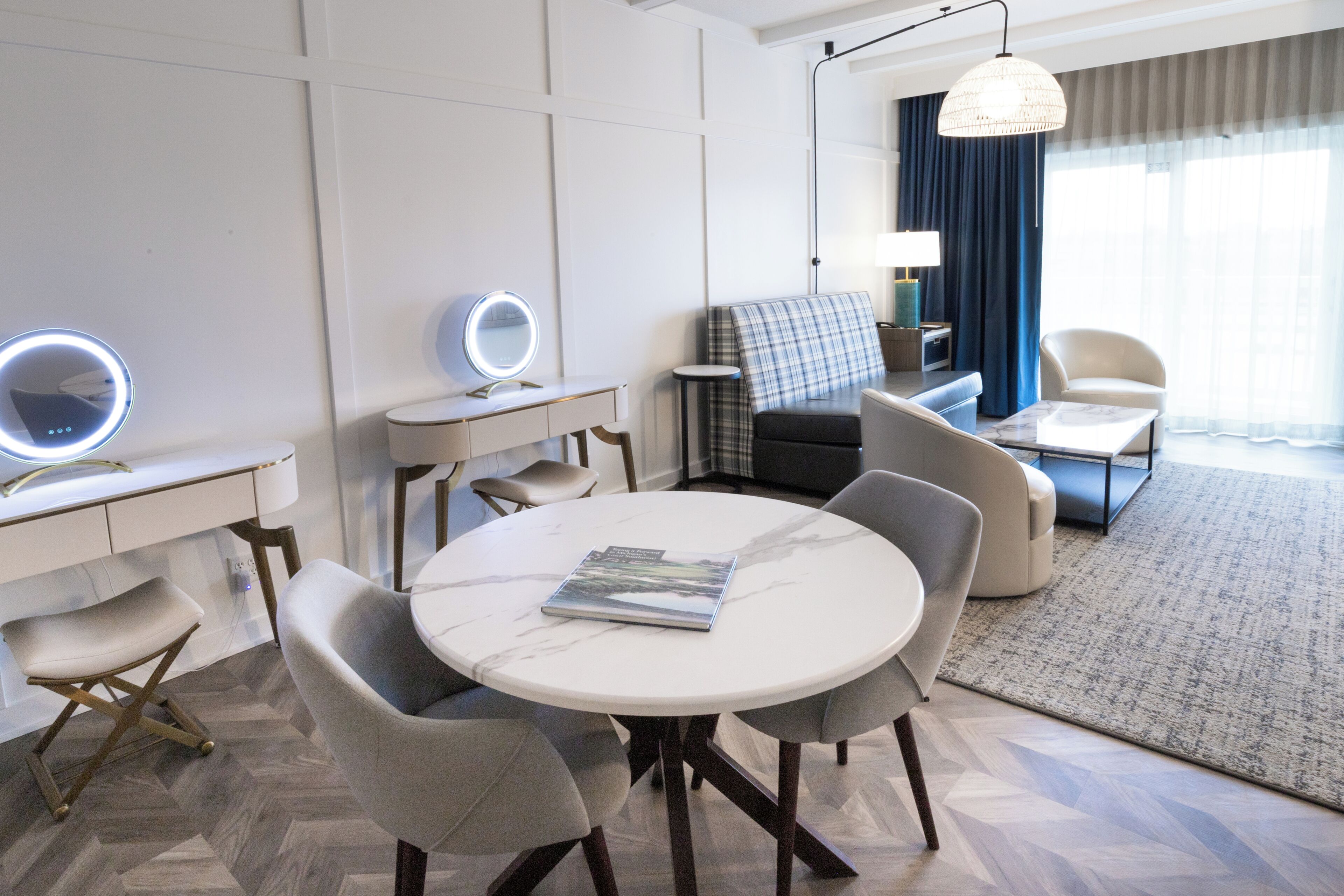 A modern hotel room with a round dining table, a sofa, a chair, a small side table, and a lamp.