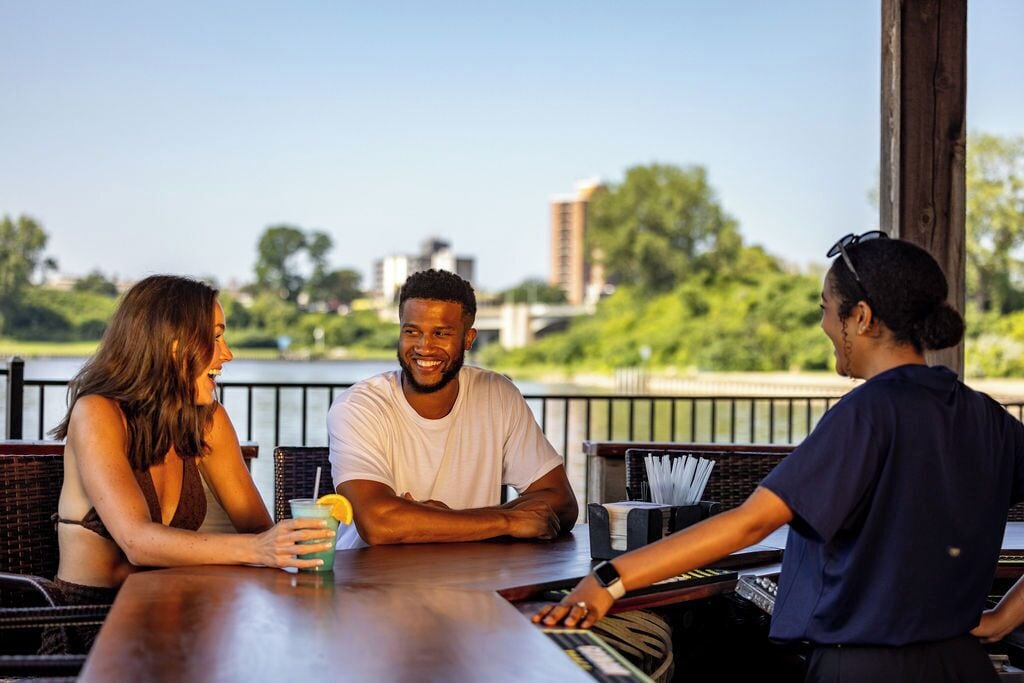 A group of people seated at an outdoor table with a view of a body of water and buildings in the background.