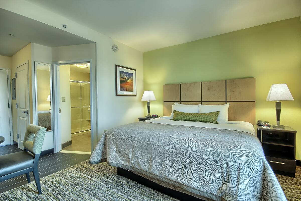 A hotel room with a large bed, two bedside lamps, a headboard, a framed picture on the wall, and a chair by the window.