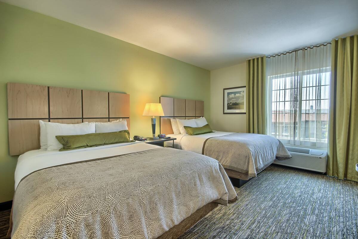 A hotel room with two beds, a nightstand, a lamp, a window with curtains, and a framed picture on the wall.