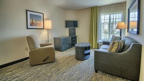 A modern hotel room with a sofa, armchair, and a small round table. There is a flat-screen TV mounted on the wall, a lamp, and a framed picture on the wall. - Candlewood Suites Columbus - Grove City by IHG (Grove City)