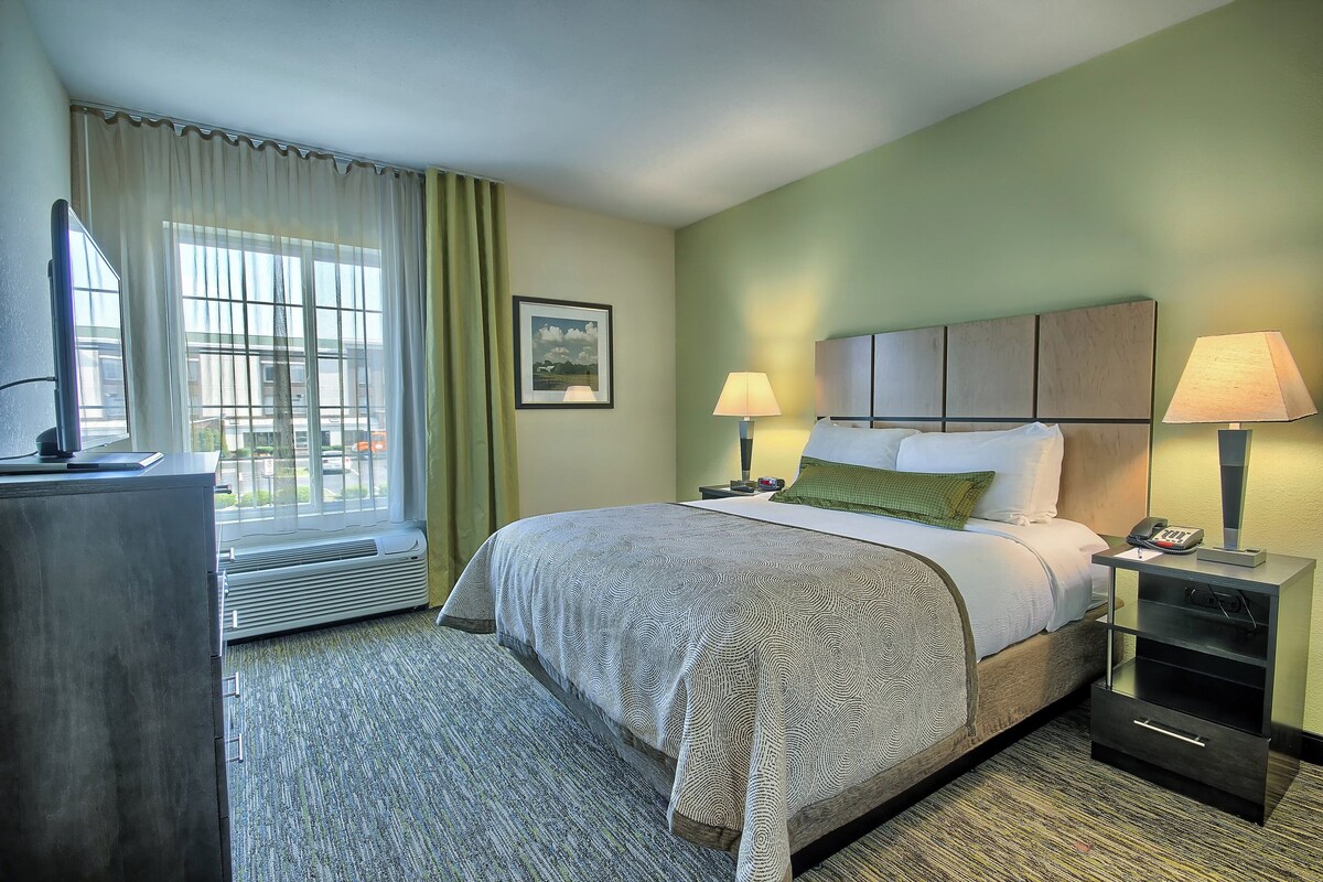 Ein Hotelzimmer mit einem Bett, zwei Nachttischlampen, einem Nachttisch, einem Schreibtisch mit Stuhl, einem Fenster mit Vorhängen und einem gerahmten Bild an der Wand.