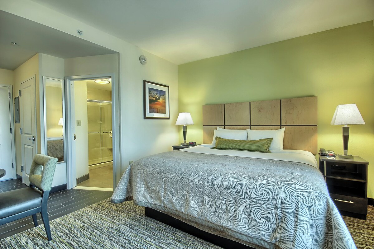 Ein Hotelzimmer mit Bett, zwei Nachttischlampen, einem Nachttisch, einem Sessel, einer Tür zum Badezimmer und einem gerahmten Bild an der Wand.