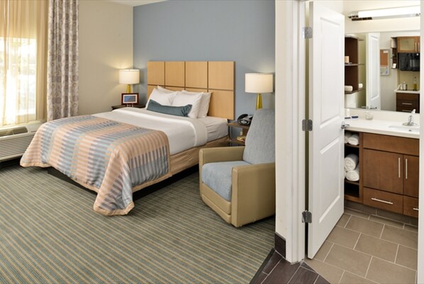 Une chambre d’hôtel comprenant un lit, une chaise, une table de chevet, une lampe, une salle de bain avec un lavabo et un miroir, et une fenêtre avec des rideaux.