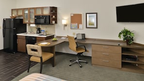 Une chambre d’hôtel compacte comprenant un coin cuisine, un bureau avec une chaise, un téléviseur à écran plat et une plante en pot.