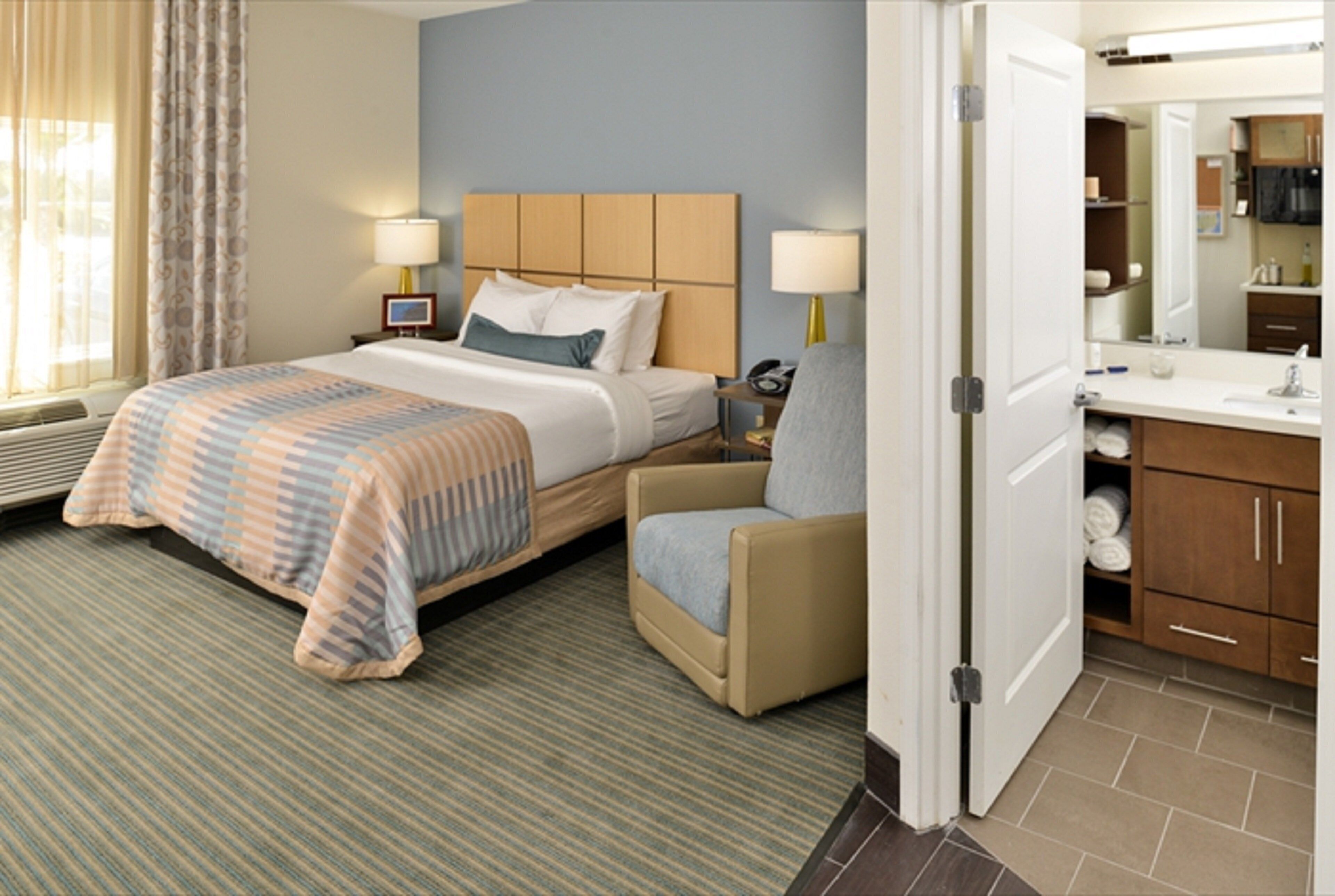 Une chambre d’hôtel comprenant un lit, une chaise, une table de chevet, une lampe, un miroir et une salle de bain avec un lavabo et des placards.
