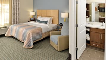 A hotel room with a bed, a chair, a nightstand, a lamp, a mirror, and a bathroom with a sink and cabinets.