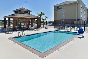 A swimming pool with lounge chairs and a covered seating area, surrounded by a fence.