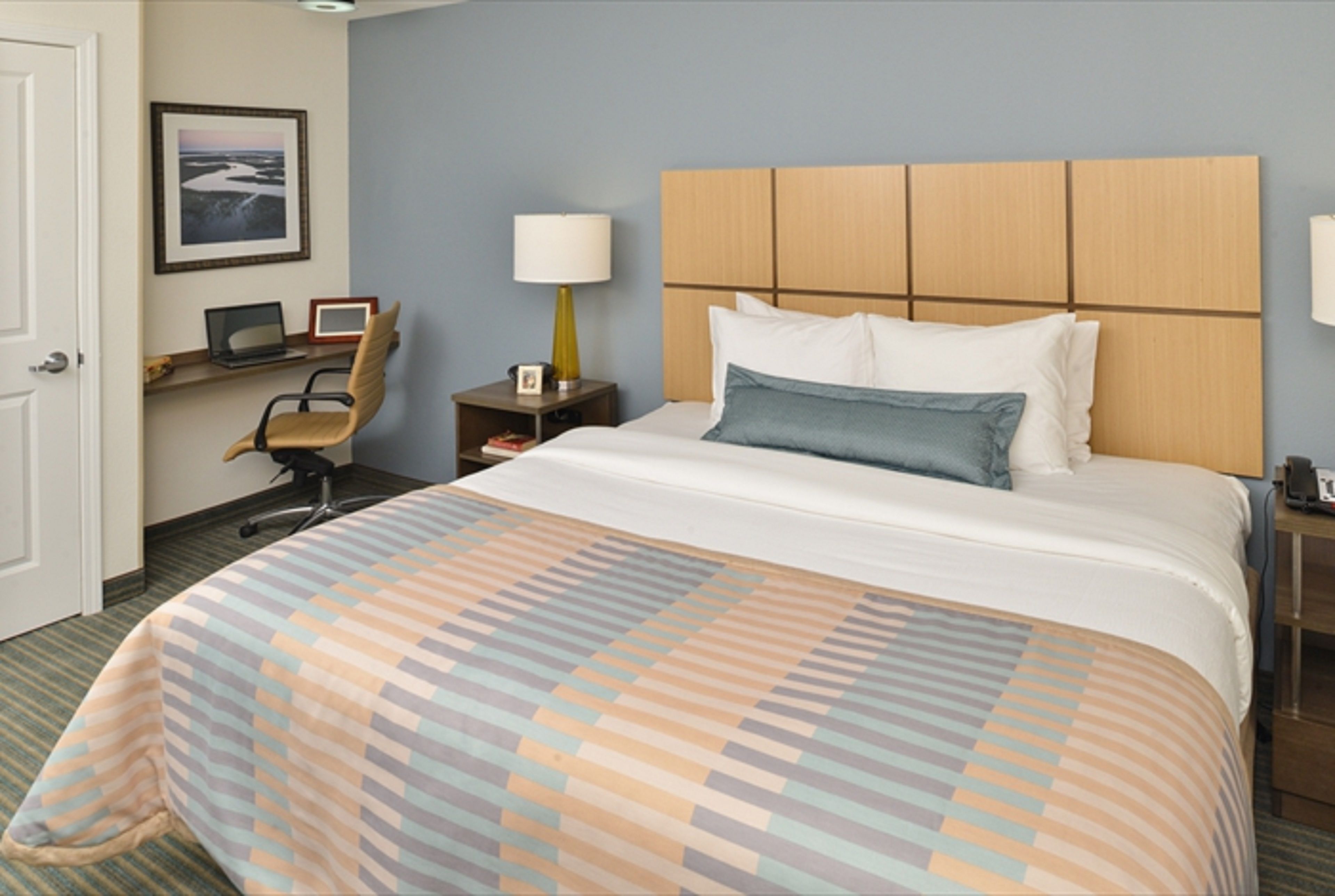 Une chambre d’hôtel avec un lit, un bureau avec un ordinateur portable, une chaise, une lampe et un tableau encadré au mur.
