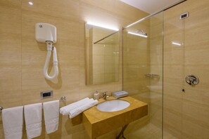 Un baño moderno con pared de azulejos color beige, tocador de madera, lavamanos blanco, cabina de ducha de vidrio y un perchero con toallas blancas.
