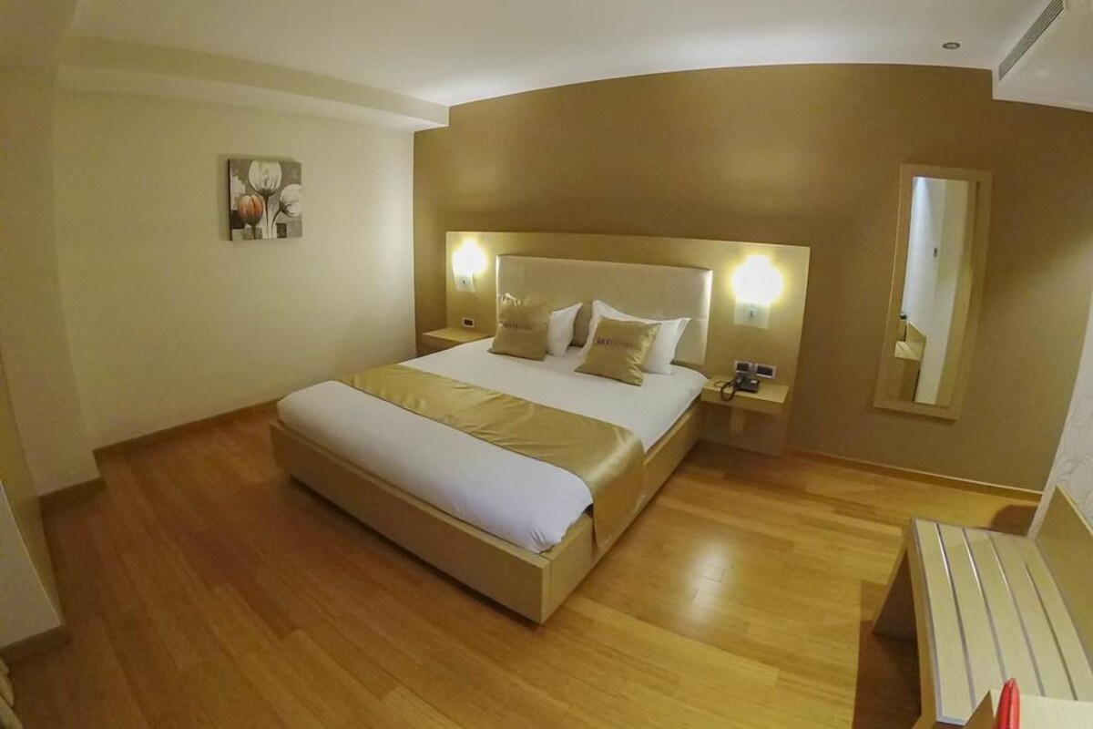 A hotel room with a large bed, wooden flooring, a nightstand, a bedside table, and a mirror.