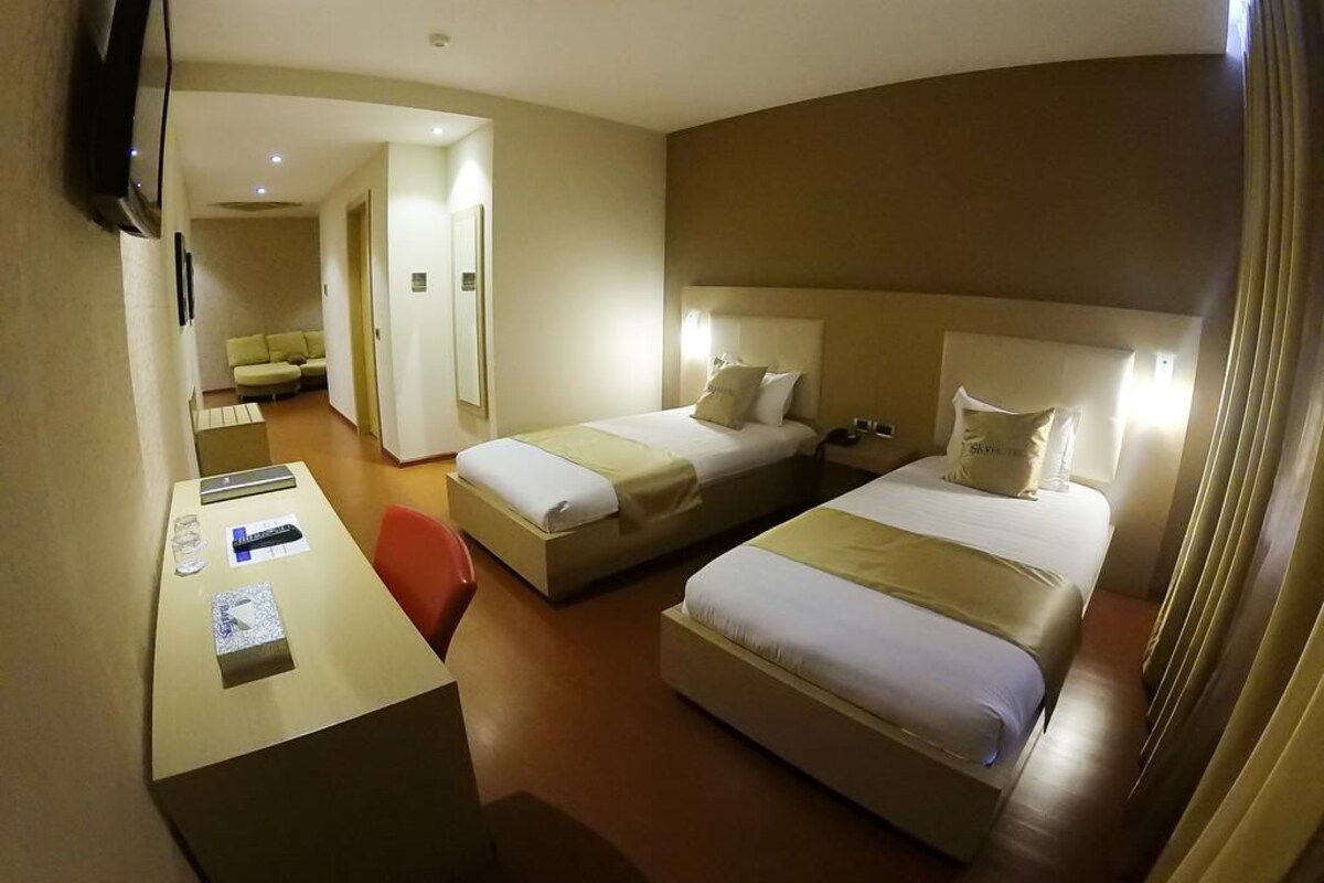 A hotel room with two beds, a desk, and a chair. There is a sofa in the background.