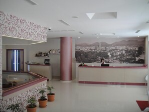 A hotel reception area with a large cityscape mural, a reception desk, and a glass partition with potted plants. - Sky 2 (Tirana)