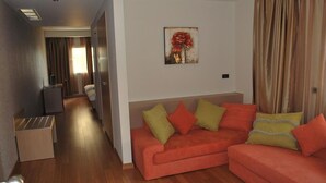 A hotel room with a wooden floor, a red sofa with multiple cushions, a painting on the wall, and a curtain on the window.