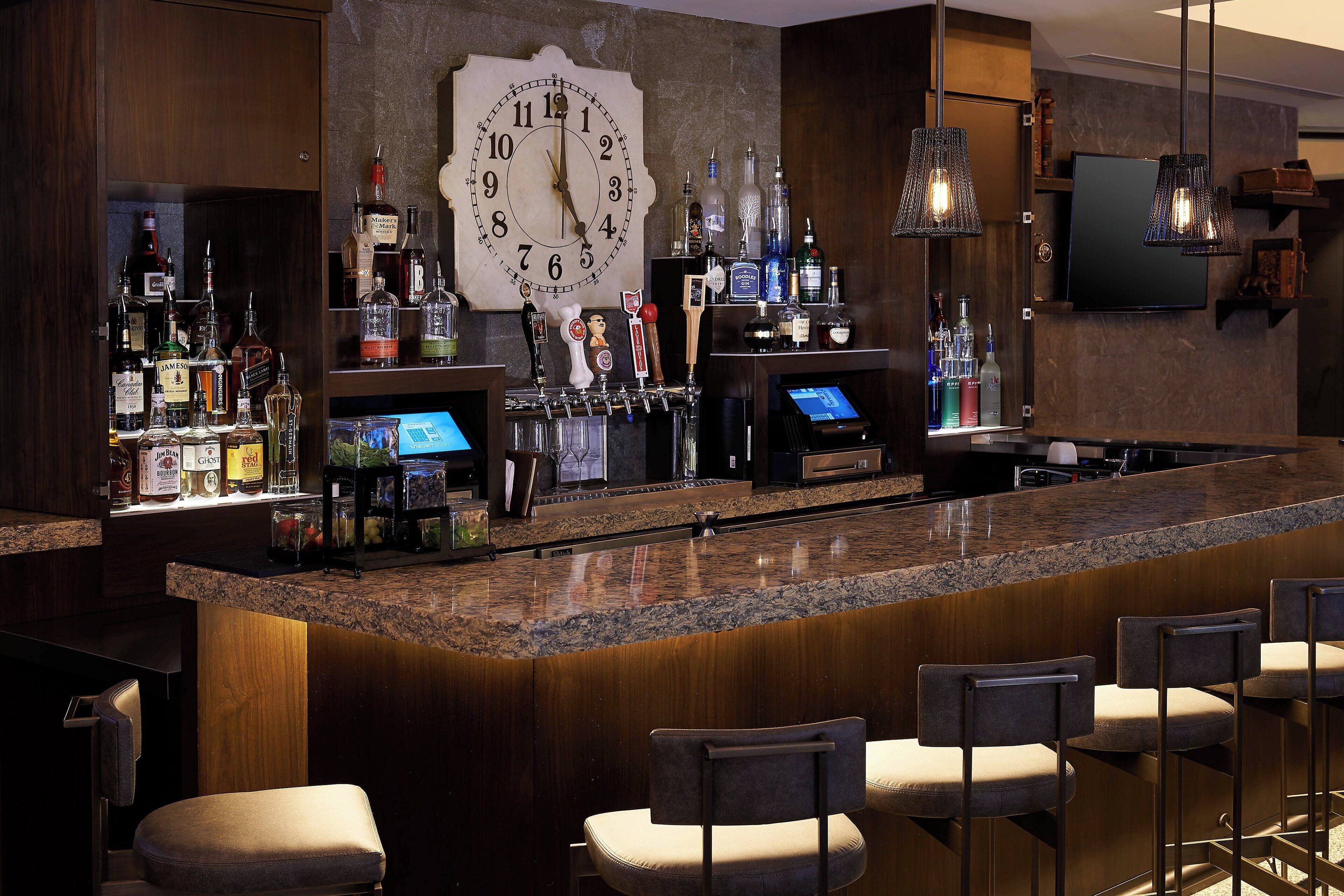 A bar area with a large clock, bar stools, and a variety of bottles.
