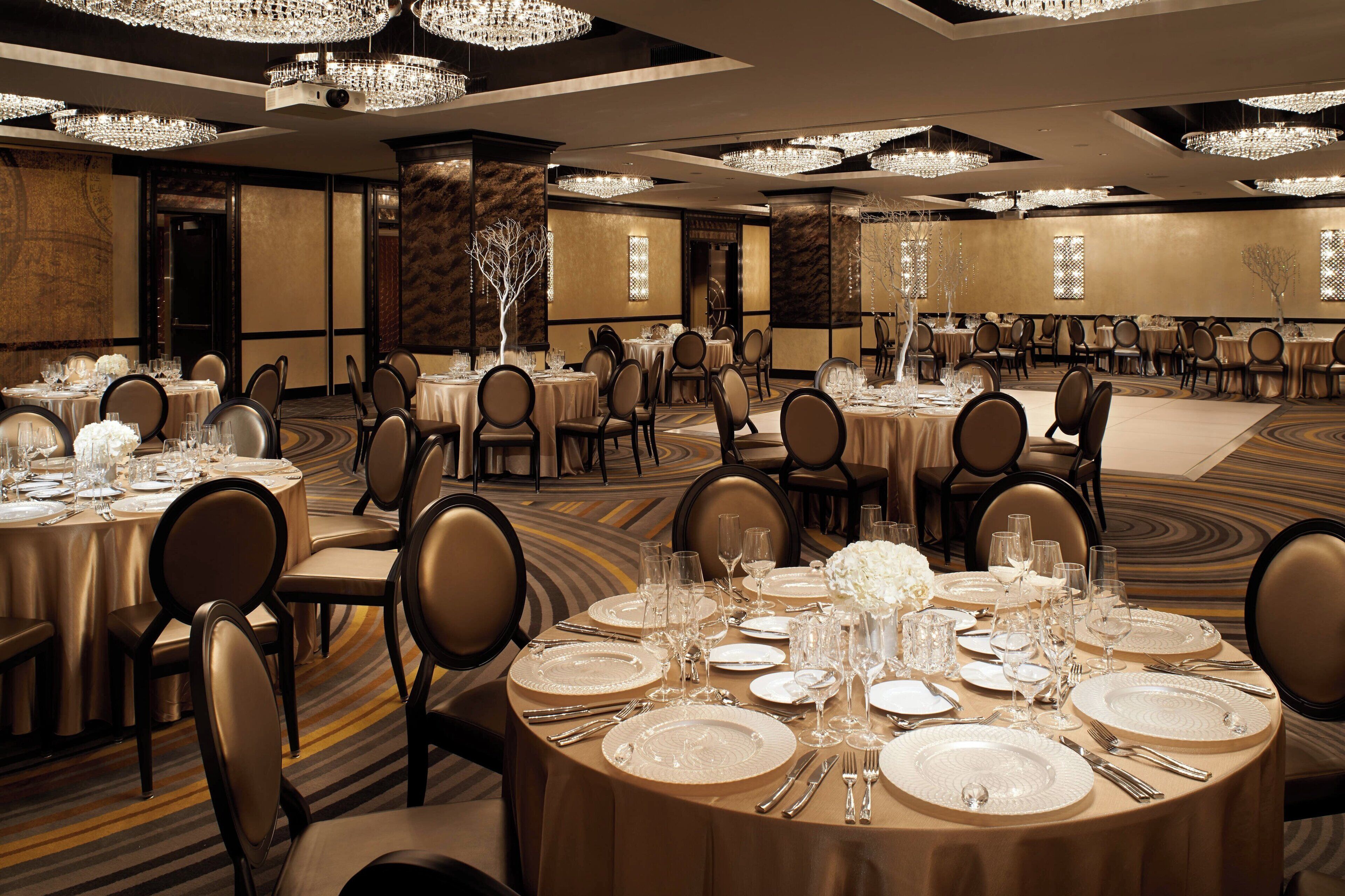 A banquet hall with round tables set for dining, featuring chandeliers and a carpeted floor.