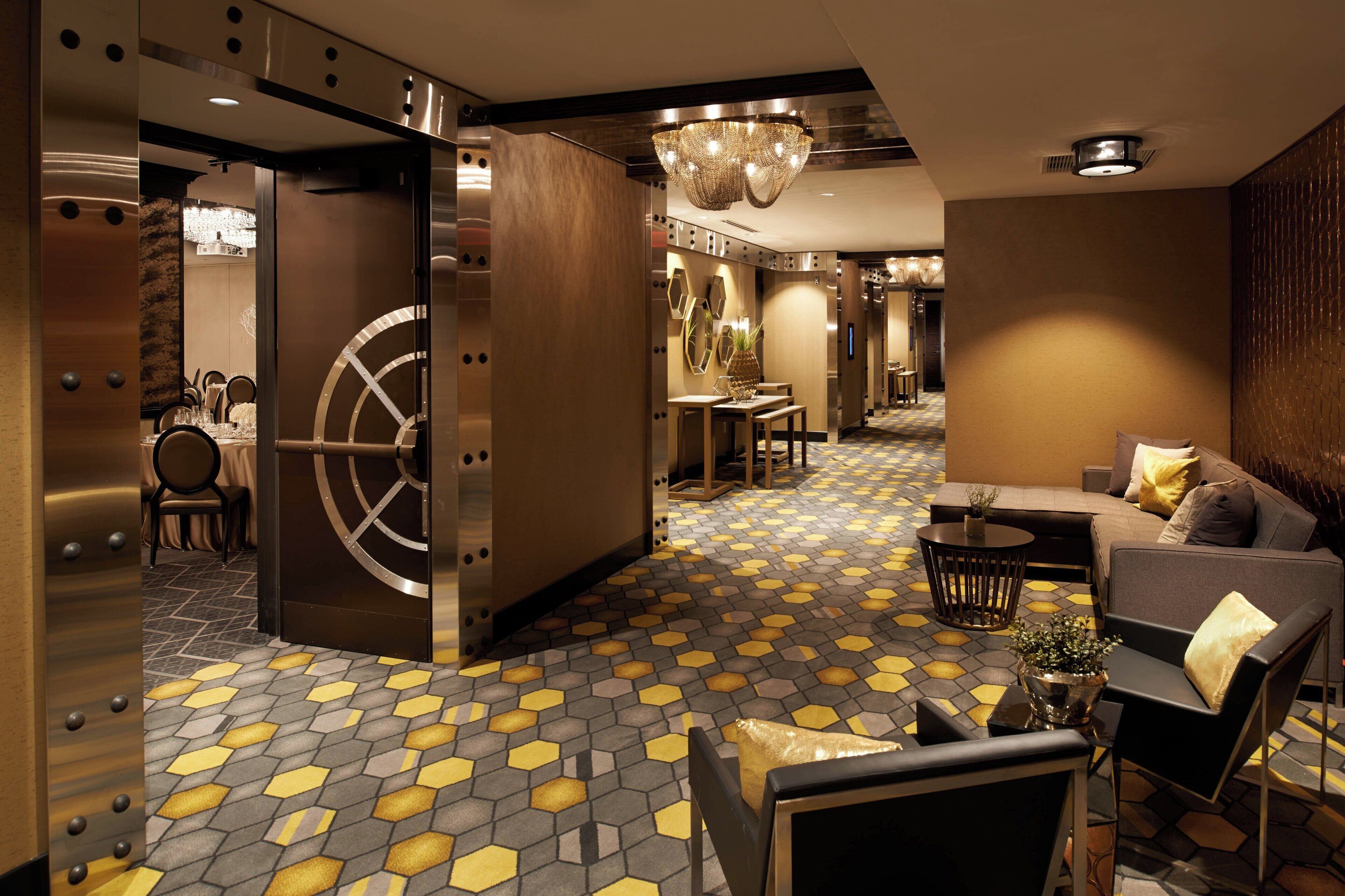A modern hotel lobby with a geometric patterned floor, a large safe, and a cozy seating area with a chandelier.
