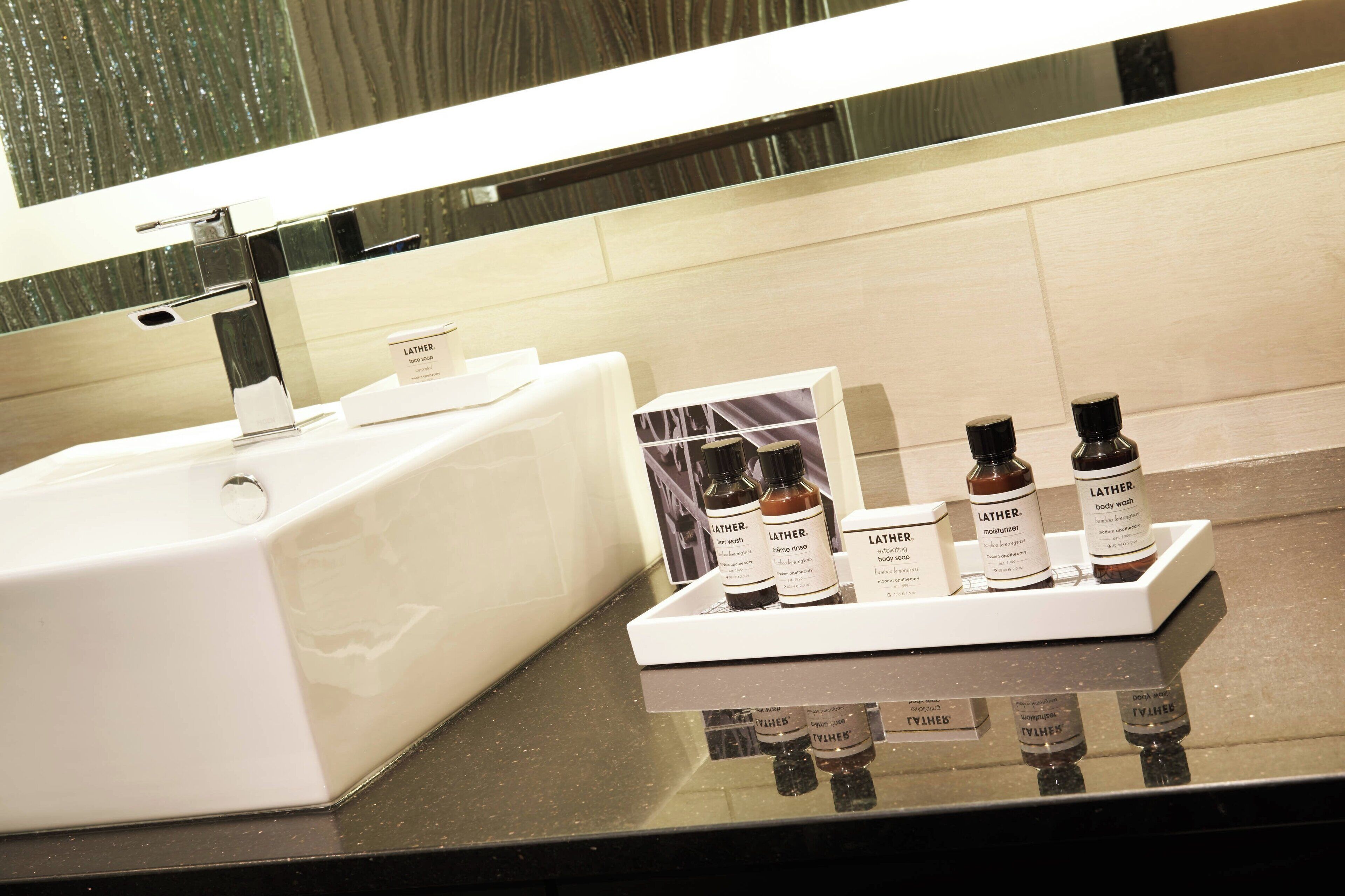 Casa de banho com lavatório branco e espelho, um tabuleiro com várias embalagens de gel de banho Lather e um dispensador de sabonete branco.