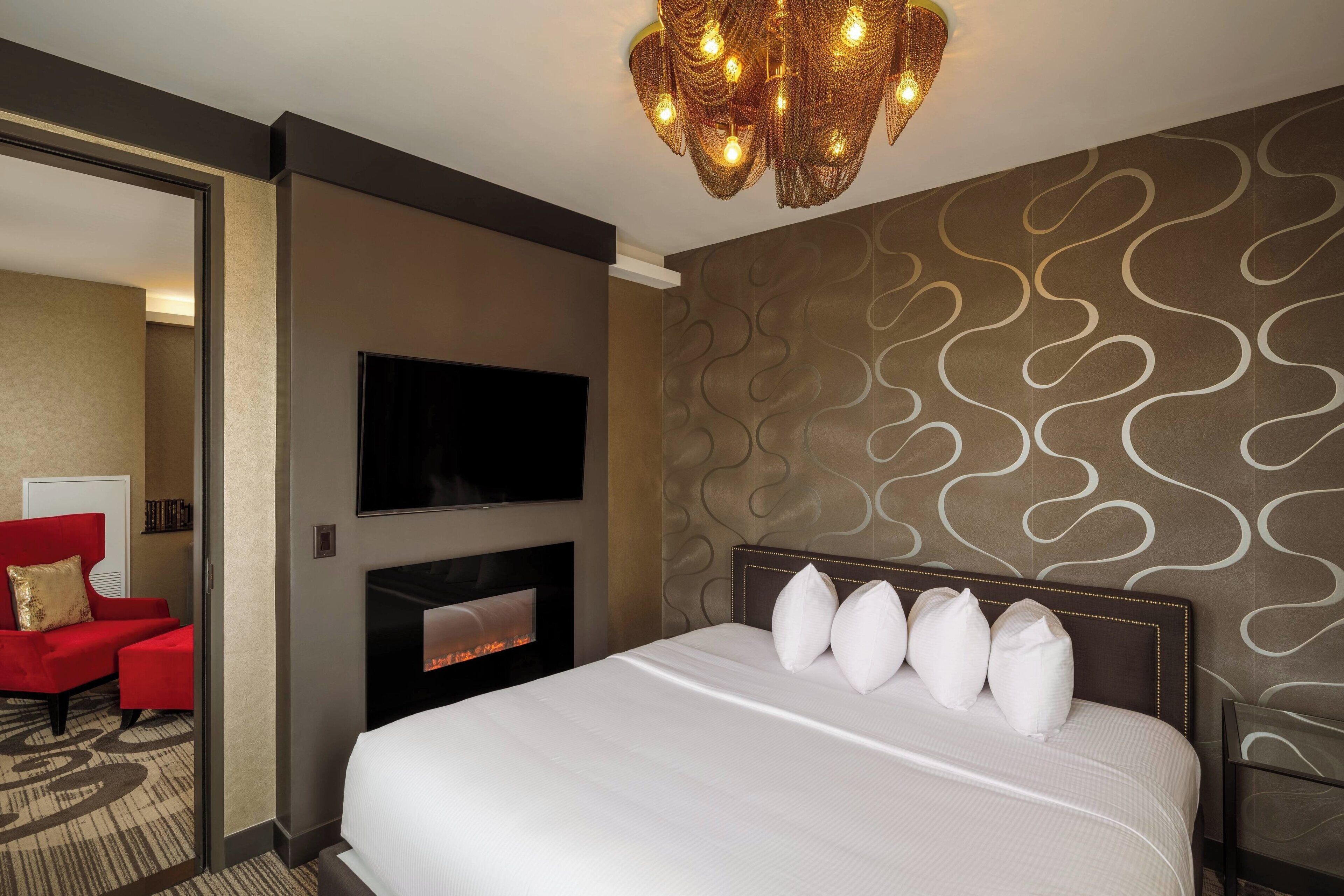 A hotel room with a large bed, a flat-screen TV, a fireplace, and a red armchair.