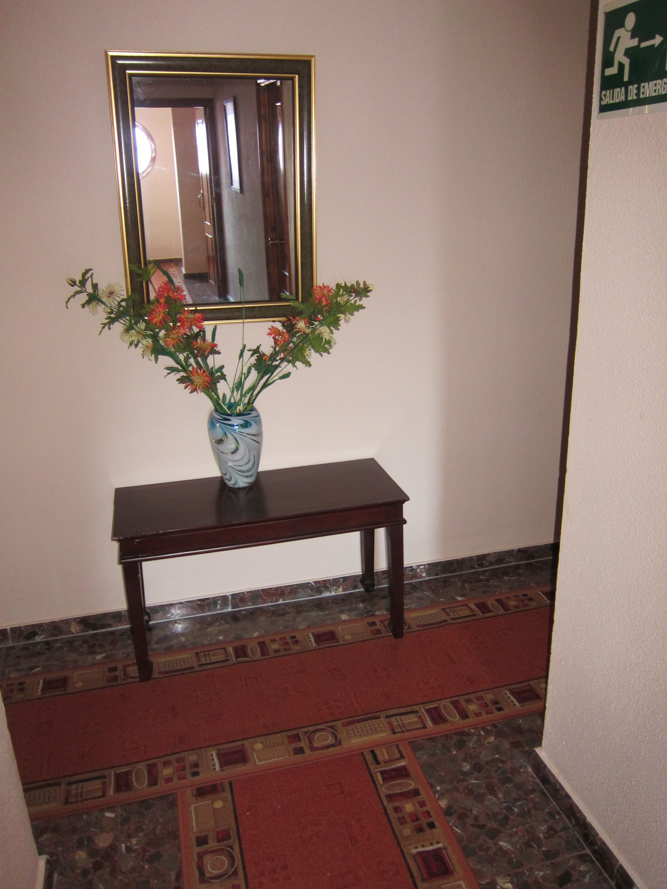 Un pasillo con un espejo en la pared, un jarrón con flores sobre una mesa y piso alfombrado.
