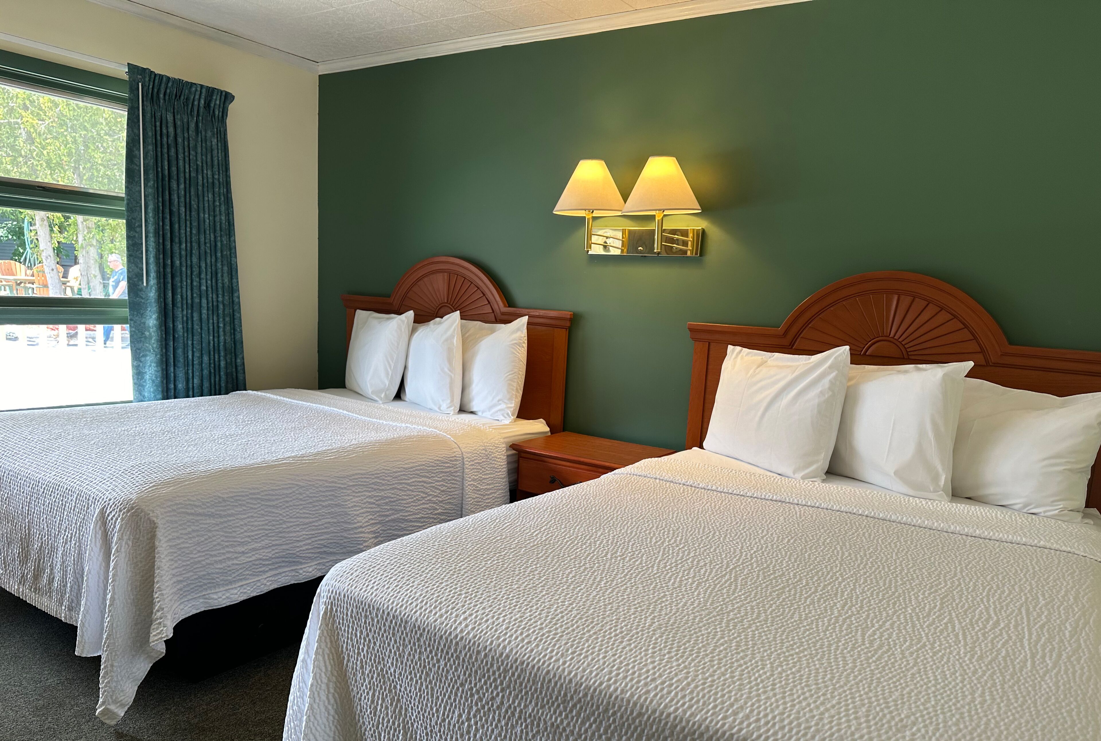 A hotel room with two beds, a wooden headboard, a nightstand, and a wall-mounted lamp.