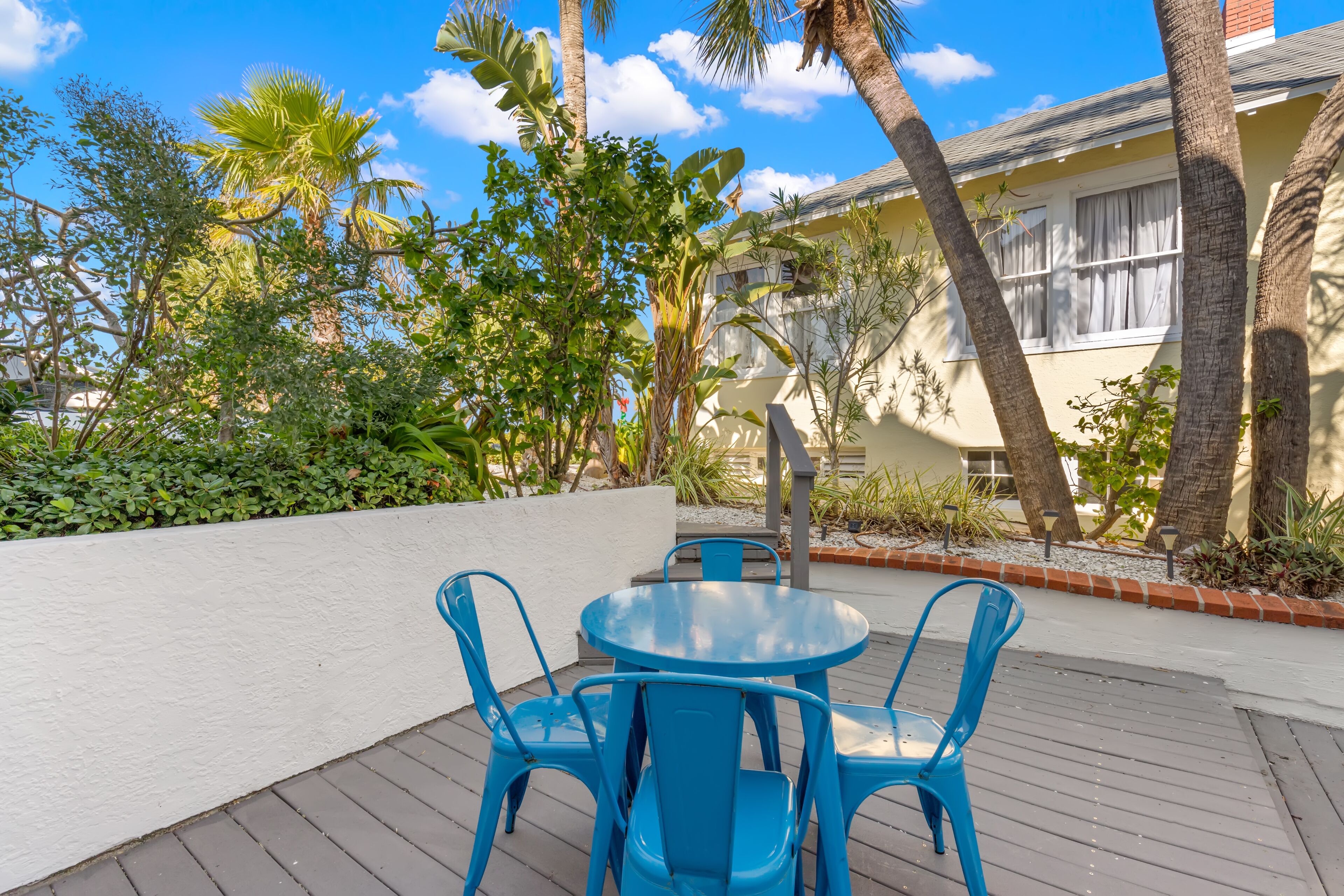 A patio with a round table and blue chairs, surrounded by tropical plants and a building with a white exterior.