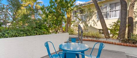 A patio with a round table and blue chairs, surrounded by tropical plants and a building with a white exterior.