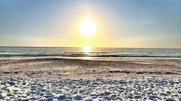 Uma praia com pegadas a levar à água, céu claro e o sol a pôr-se ou a nascer no horizonte.