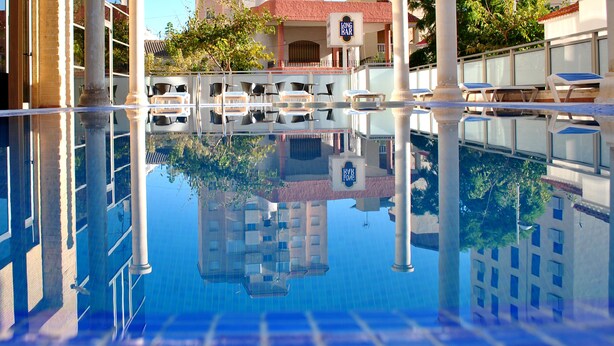 A clear view of a swimming pool with reflections of surrounding buildings and a bar area. - Hotel Biarritz (Gandia)