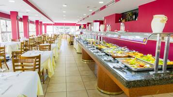 Un'area buffet con diverse postazioni di cibo, tavoli e sedie, e interni luminosi con pareti rosa e ampie finestre.
