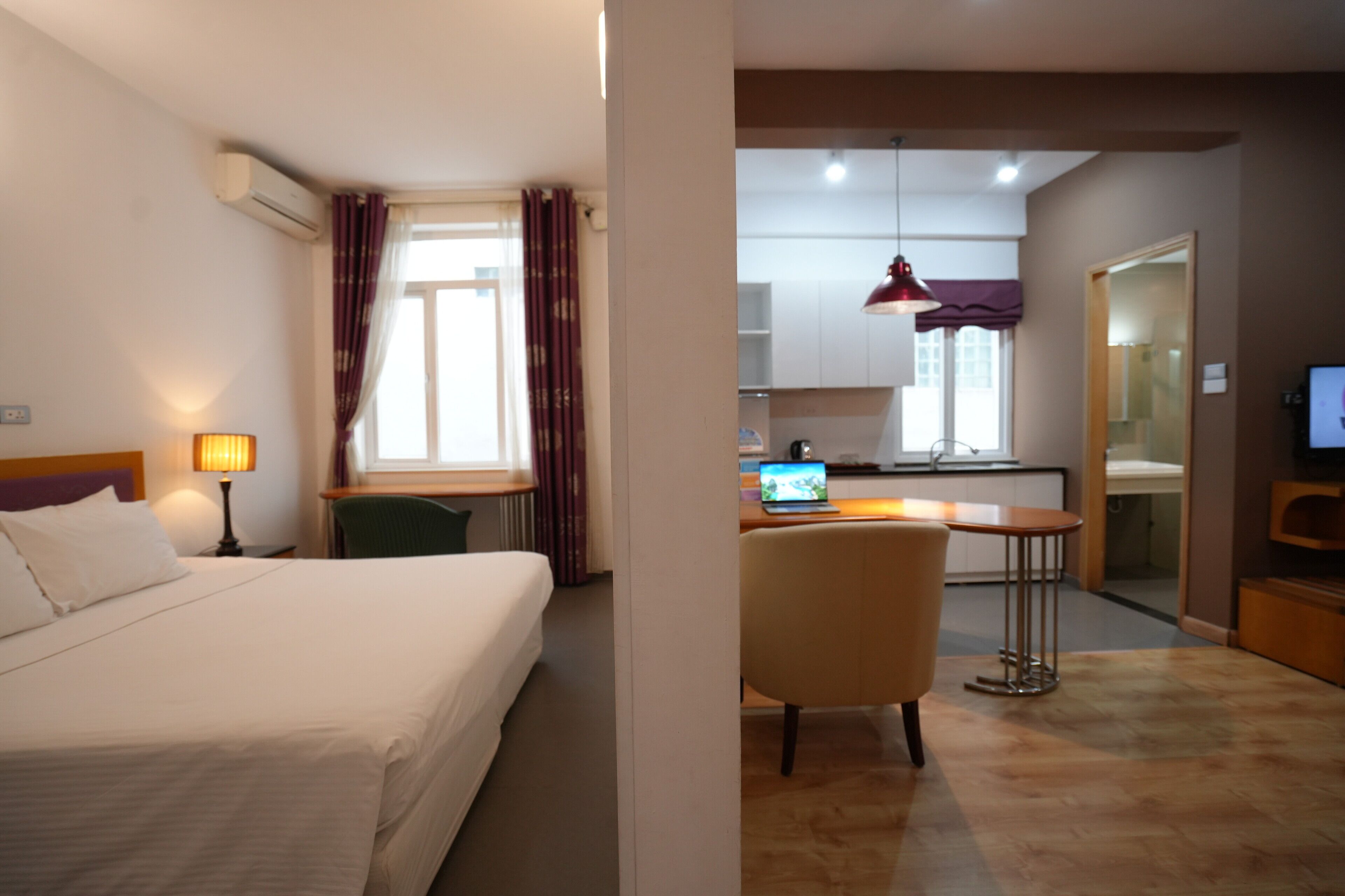 Quarto de hotel com uma cama, uma mesa com um laptop, uma cadeira, uma TV e uma cozinha ao fundo.