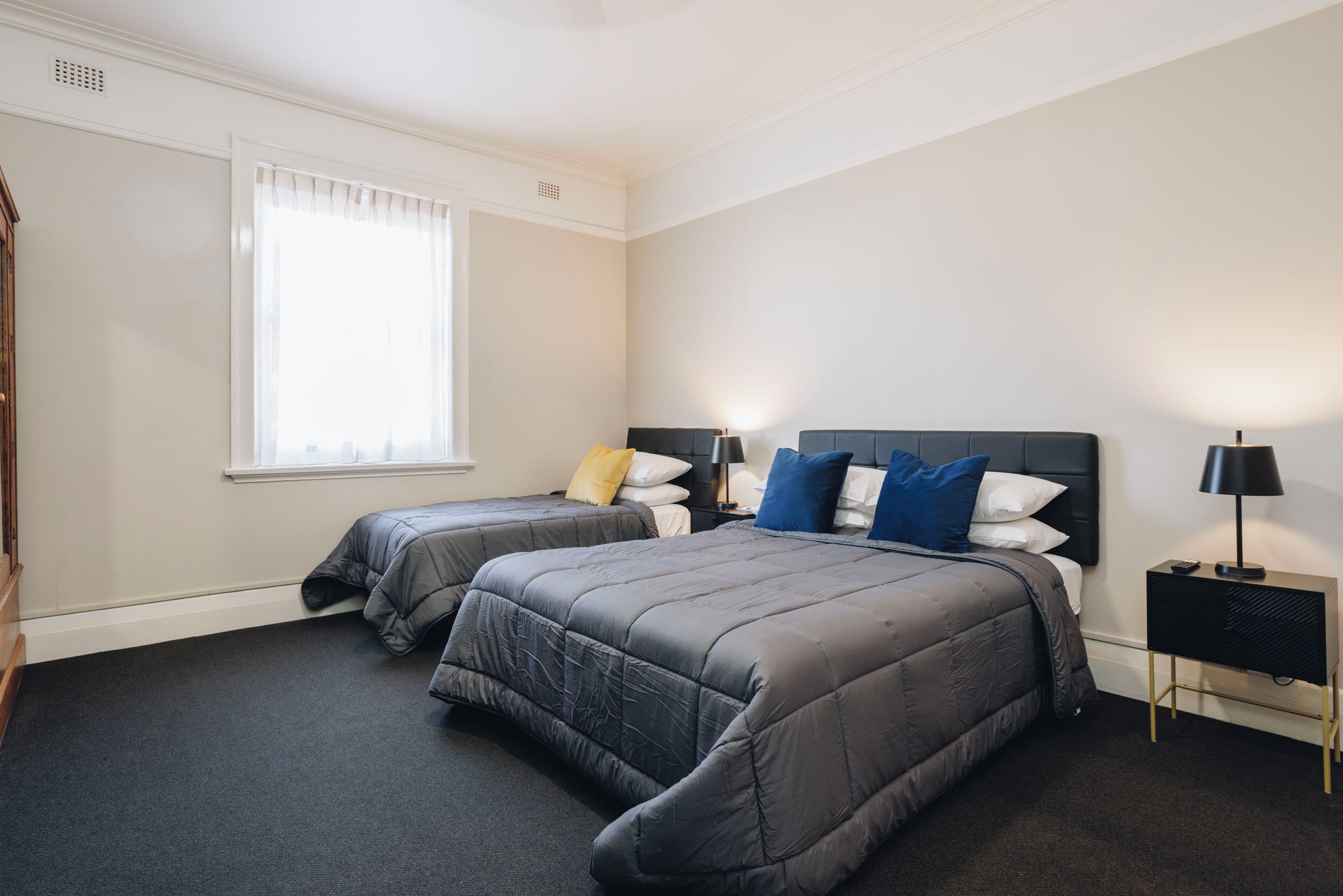 A hotel room with two beds, a window with curtains, a nightstand with a lamp, and a wall-mounted heater.