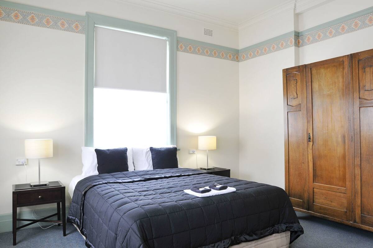 A bedroom with a bed, two bedside tables with lamps, a wardrobe, and a window with blinds.