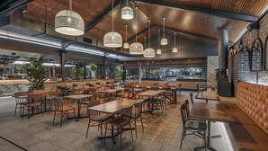 A spacious indoor dining area with multiple wooden tables and chairs, hanging pendant lights, and a brick wall with a kitchen in the background. - Hotel Gosford (Gosford)